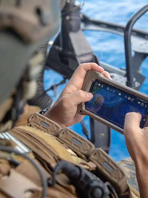 A U.S. Marine in a helicopter over open water, holding a rugged phone running a tactical map.