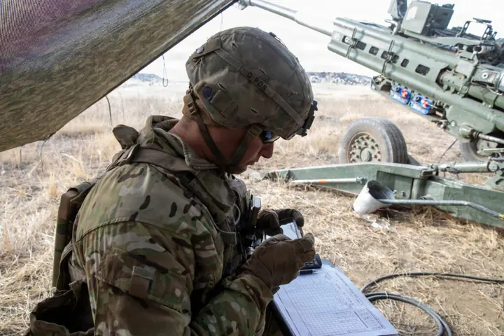 A soldier under camouflage netting reading a handheld device, a howitzer behind him.