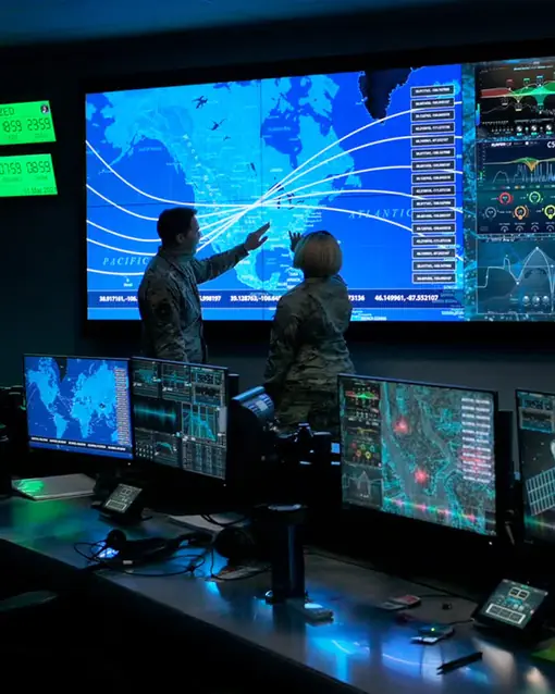 A command-center wall of live maps, two officers pointing at trajectory arcs across a continent.